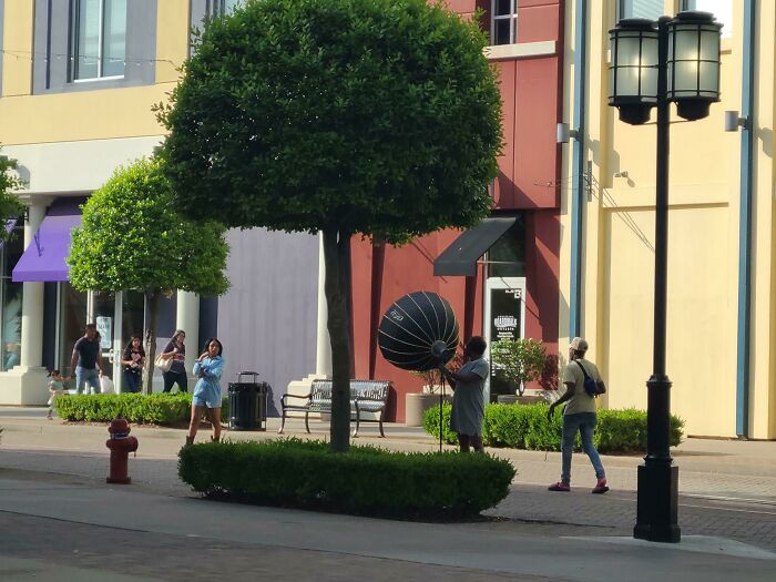 An influencer poses in a public street while an assistant holds professional lighting gear during a photo shoot.