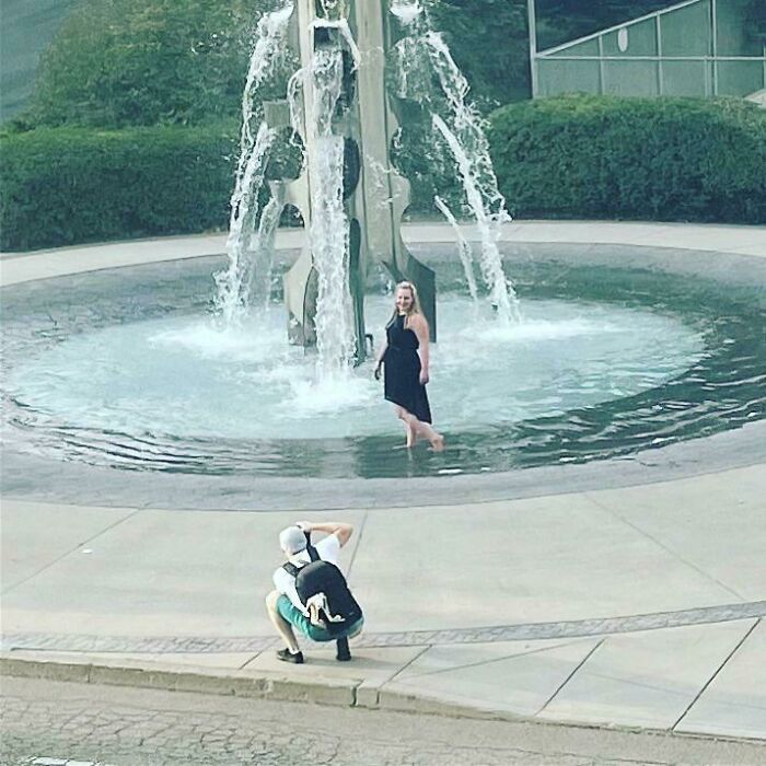 A photographer crouches in public spaces to capture an influencer posing inside a large outdoor water fountain.