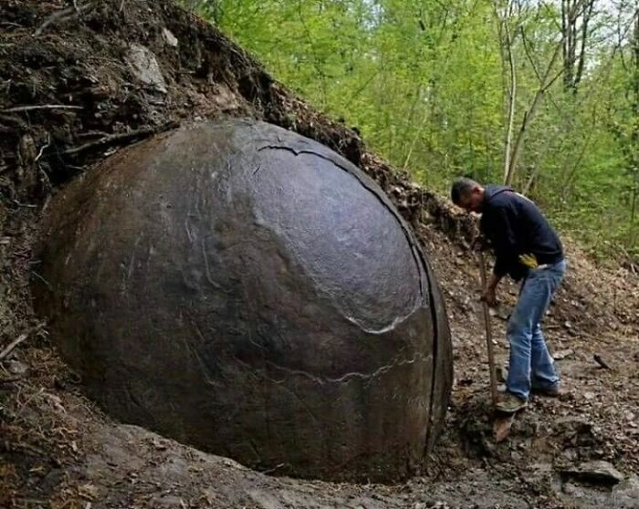 A man standing next to a massive, spherical stone embedded in the ground, showcasing surprising facts about the past.