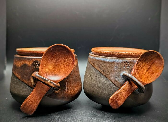 Two earthy clay pots with wooden lids and matching wooden spoons, showcasing unique pottery creations.