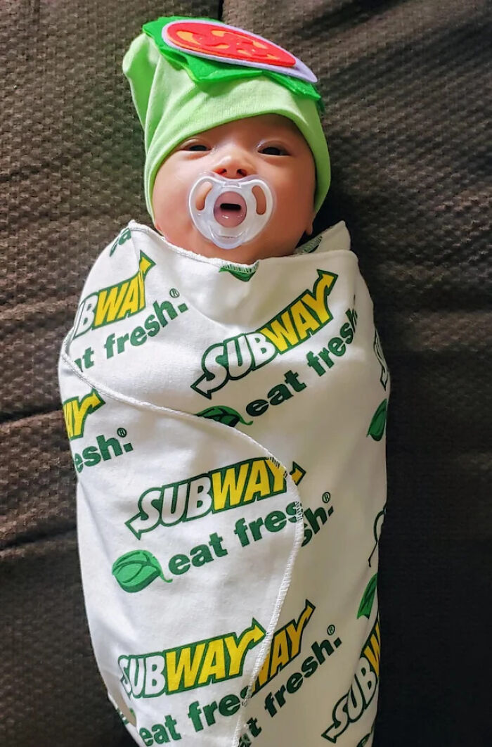 A newborn baby wrapped in a Subway sandwich swaddle blanket wearing a green hat for a funny kids Halloween costume.