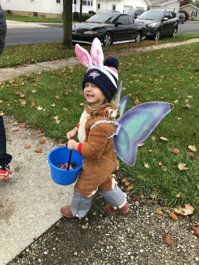 A happy toddler wearing a creative fairy bunny Halloween costume carrying a blue trick-or-treat bucket.
