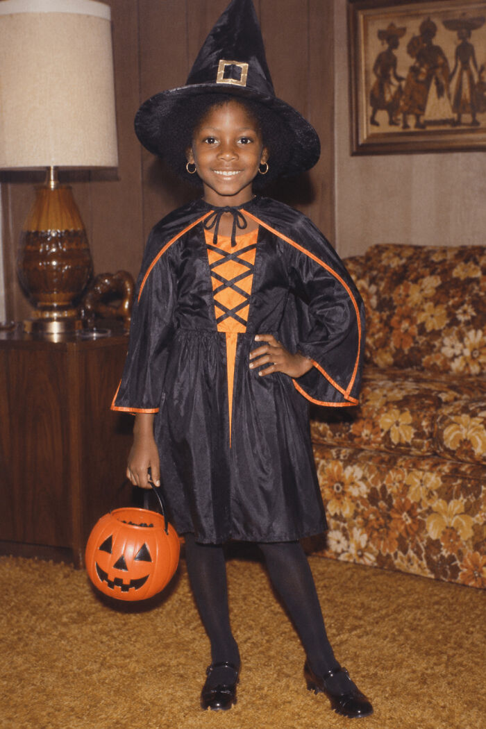 A young girl smiling in a black witch Halloween costume holding an orange pumpkin bucket