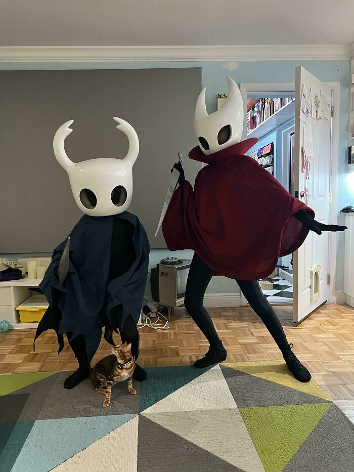 Two people wearing Hollow Knight kids Halloween costumes posing indoors with a cat