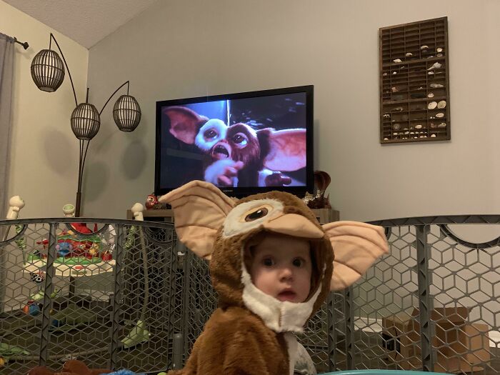 A toddler wearing a Gizmo costume sitting in front of a TV showing a Gremlins character for Halloween costumes ideas.