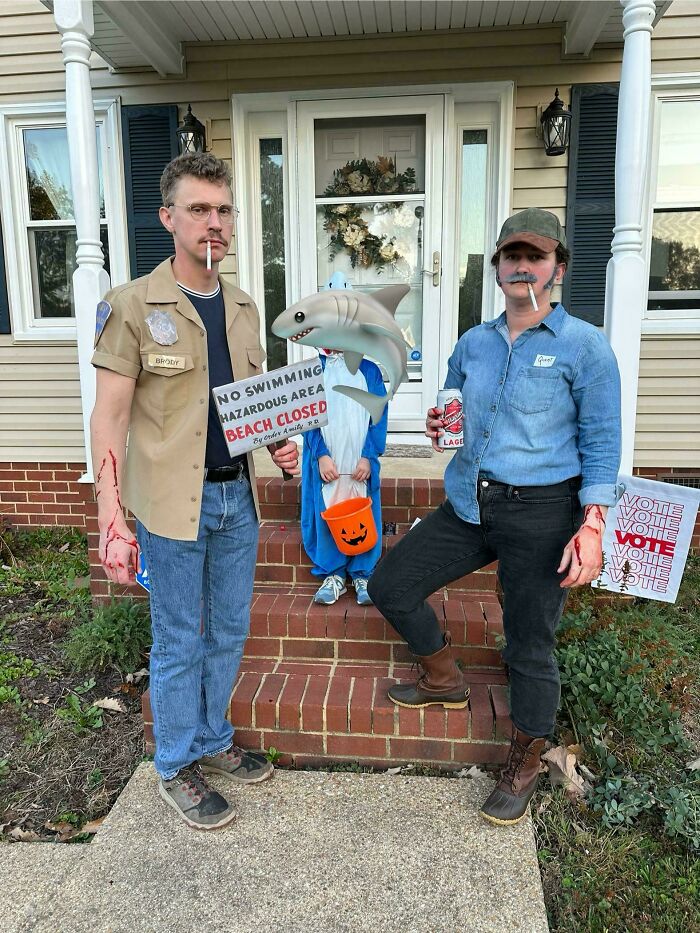 Family wearing Jaws themed Halloween costumes including a shark and beach safety officers standing on front porch steps.