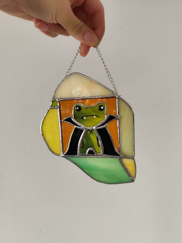 Stained glass suncatcher depicting a cute frog wearing a vampire cape for Halloween.