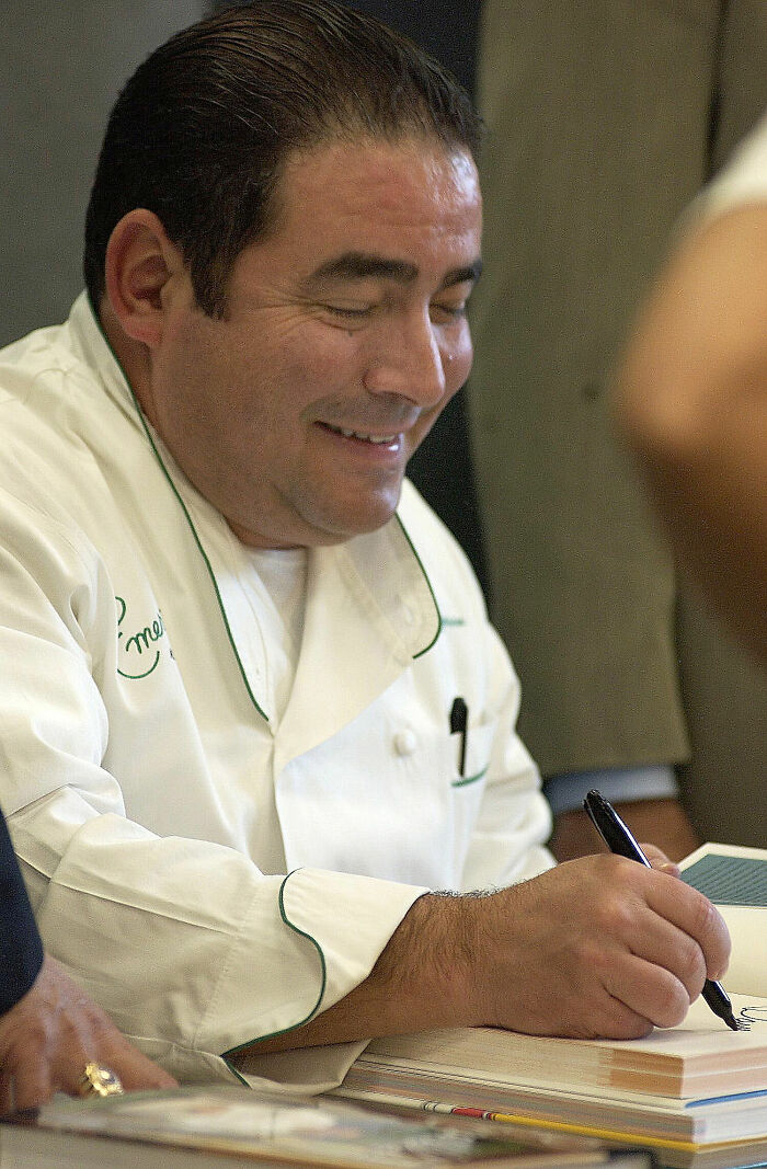 Smiling chef Emeril Lagasse wearing a white uniform and signing a book during a public appearance