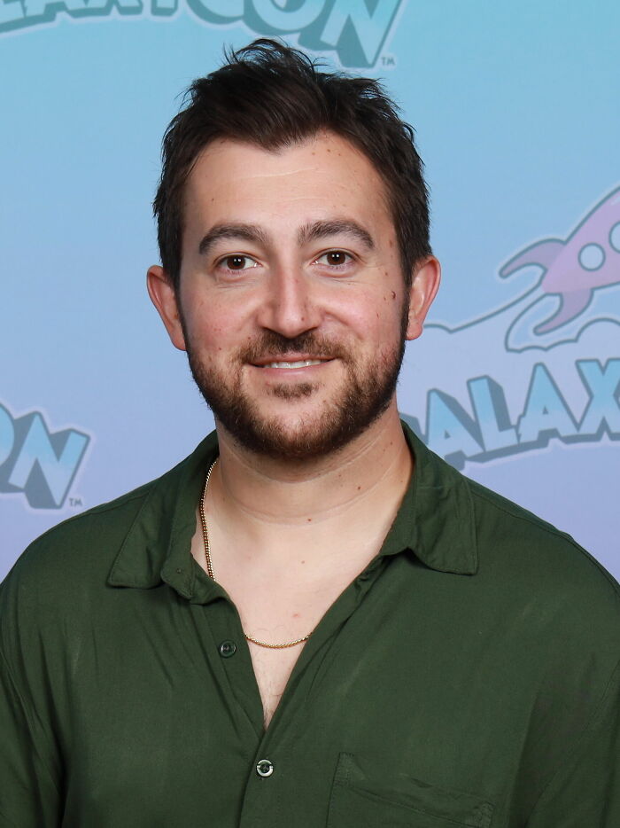 Actor Vincent Martella smiling in a green shirt against a light blue backdrop at an event