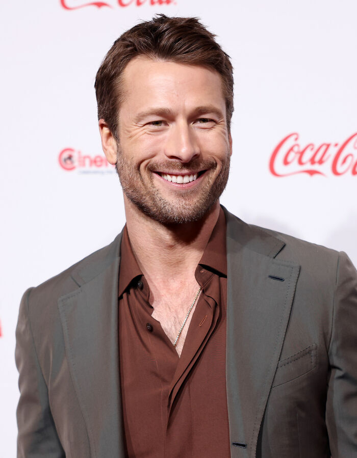 Actor Glen Powell smiling at a red carpet event in a brown shirt and suit jacket.