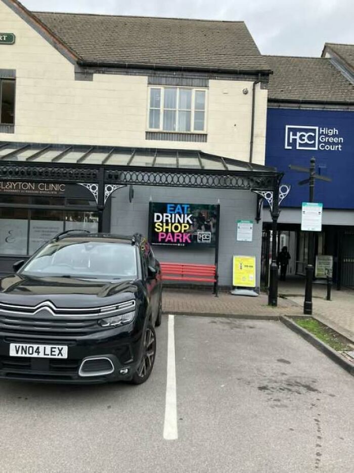 A black car parked outside a building with a funny sign featuring confusing layout instructions.