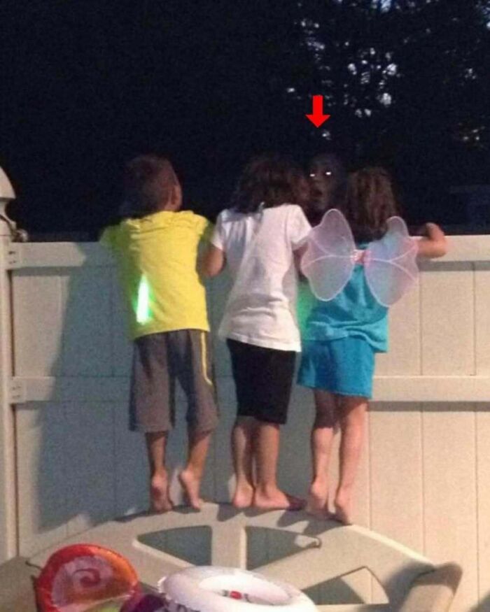 Three children look over a fence at a eerie glowing-eyed face in a real-life photo like a horror movie