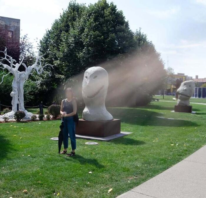 Woman standing in a park next to strange stone head sculptures, giving off horror movie vibes.