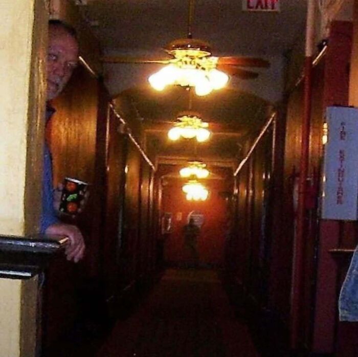 A man peering around a hallway corner with a shadowy figure behind him in a horror movie setting.