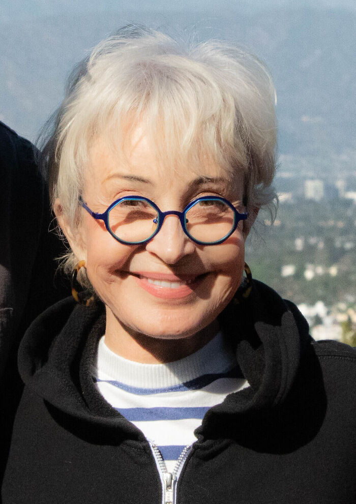 Actress Annie Potts wearing blue glasses and smiling outdoors.
