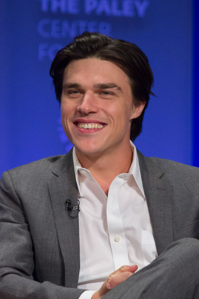 Actor Finn Wittrock smiling in a grey suit and white shirt at a panel event.