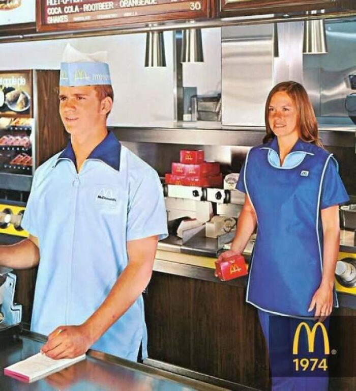 Two fast food workers wearing blue 1974 McDonald's uniforms behind the counter in old photos of America.