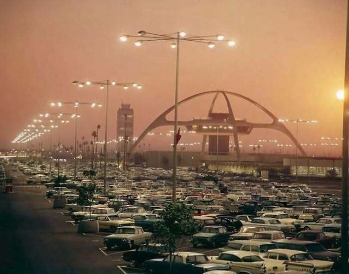Old photos of America showing a crowded parking lot full of vintage cars near an airport terminal at dusk