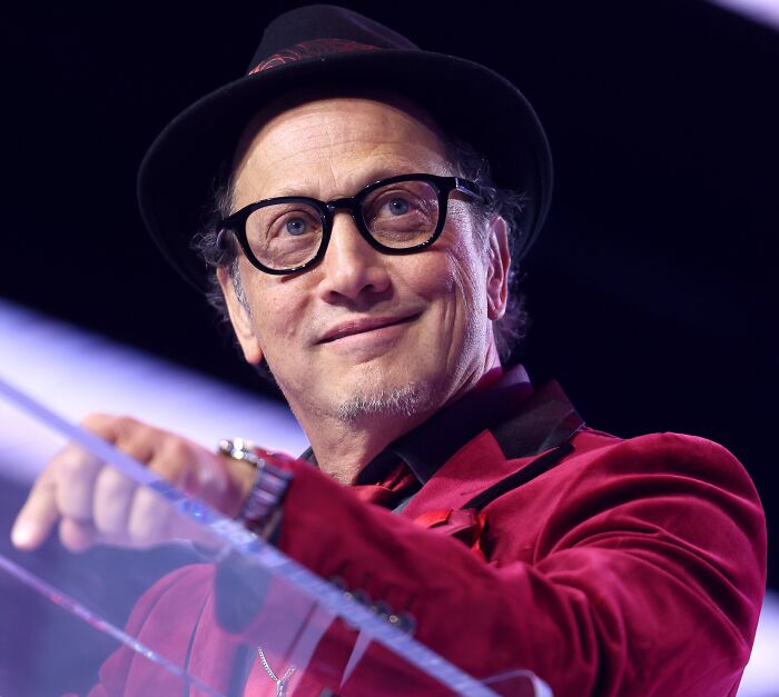 Comedian Rob Schneider wearing glasses and a hat smiling at an event.
