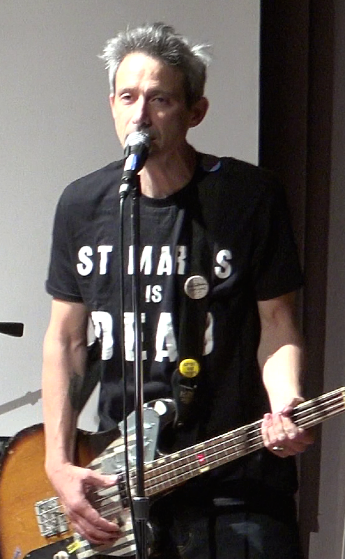 Musician Ad-Rock speaking into a microphone while playing an electric bass guitar on stage