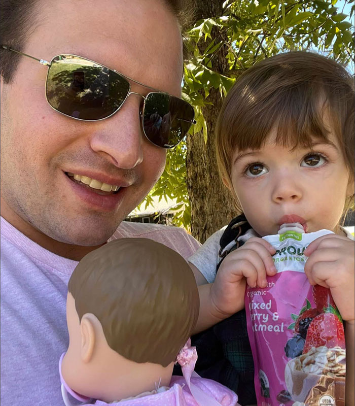 A father wearing sunglasses holding his daughter, who is drinking from a pouch A father wearing sunglasses holding his daughter, who is drinking from a pouch