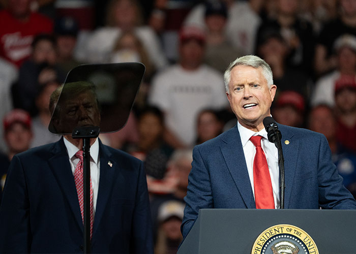 Politician speaking at a podium with Donald Trump standing nearby, amid reports of Trump waving at nobody. Politician speaking at a podium with Donald Trump standing nearby, amid reports of Trump waving at nobody.