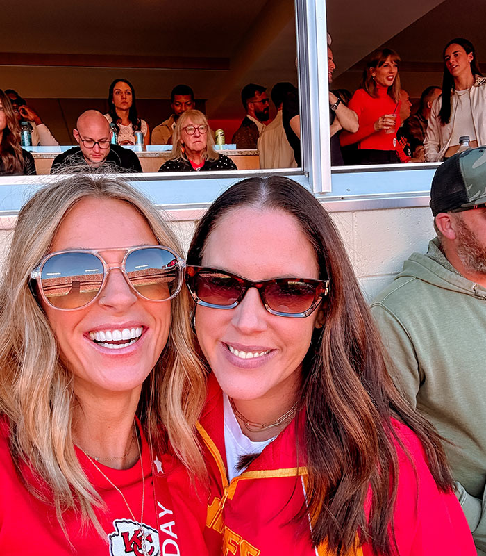 Fans posing at a Chiefs game with Taylor Swift visible in the background box