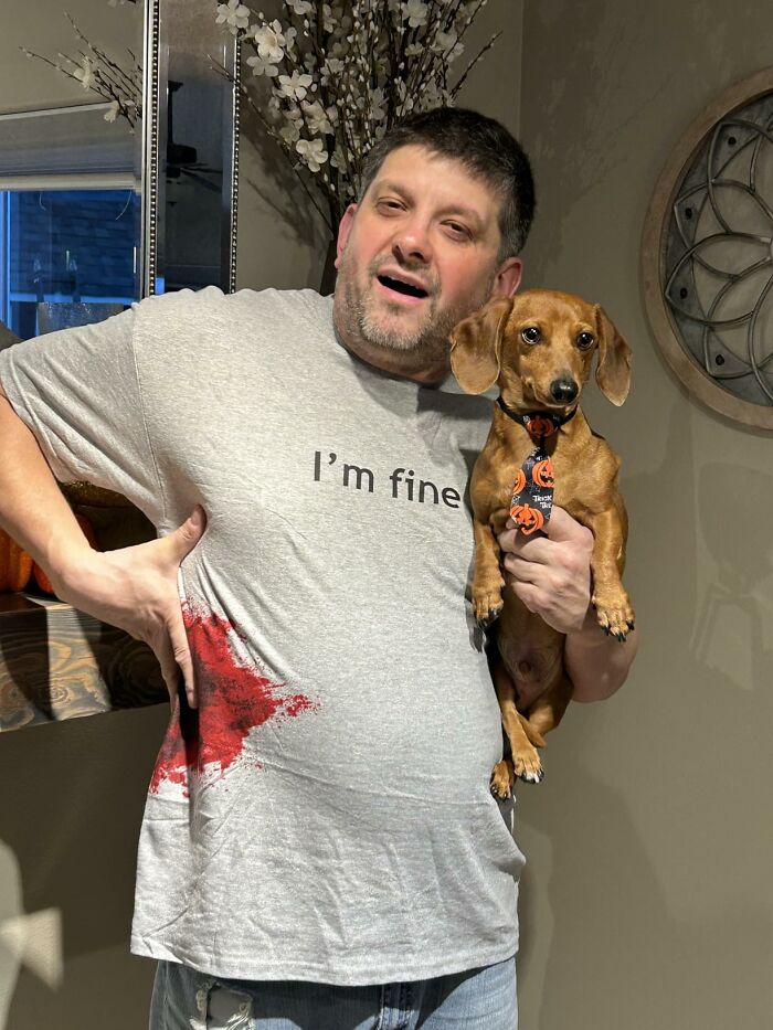A man wearing a humorous bloody I'm fine shirt holding a dachshund for a low-effort Halloween costume.
