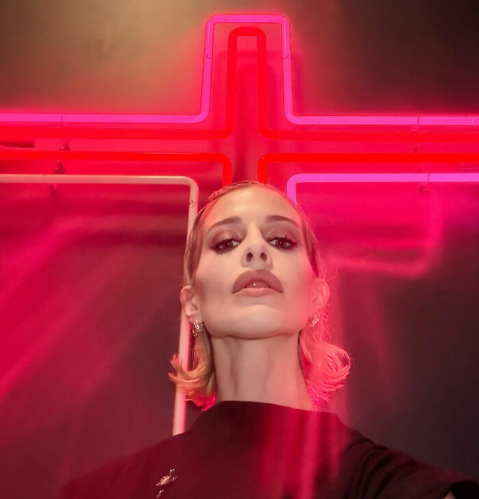 Kelly Osbourne posing beneath a bright red neon cross light.