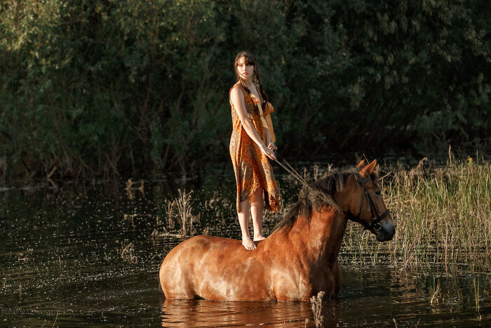 A woman standing upright on a horse in water, depicting obvious lies about equestrian skills.