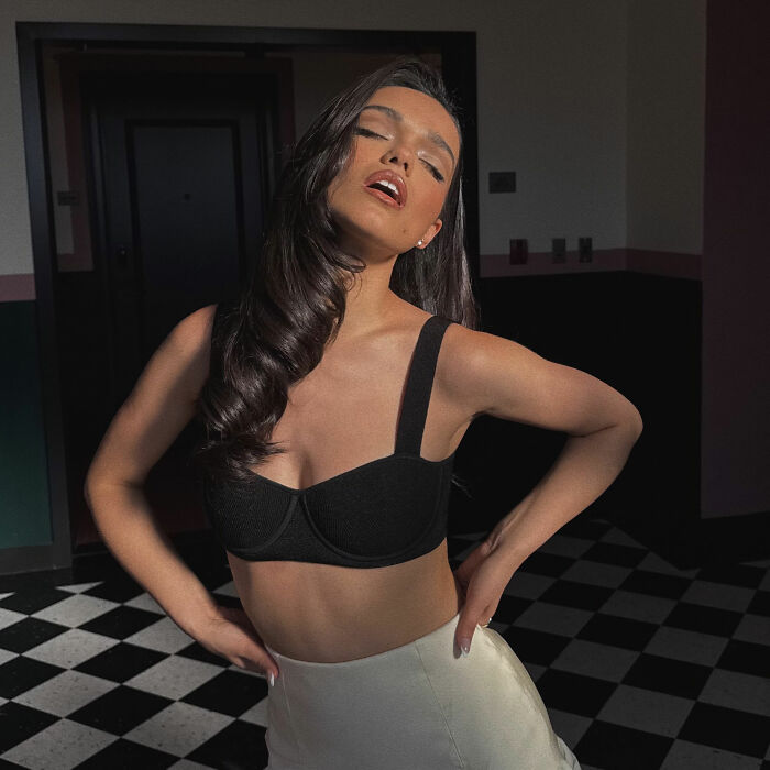 Rachel Zegler posing with closed eyes and slightly open mouth in a black crop top and white skirt.