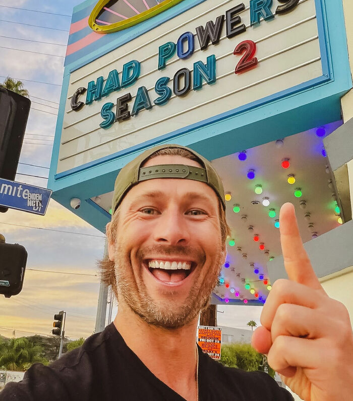 Hollywood star Glen Powell taking a selfie pointing up to a theater marquee.