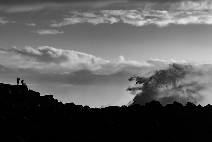 Silhouetted people standing on rocky cliffs near crashing ocean waves at a black sand Iceland coast