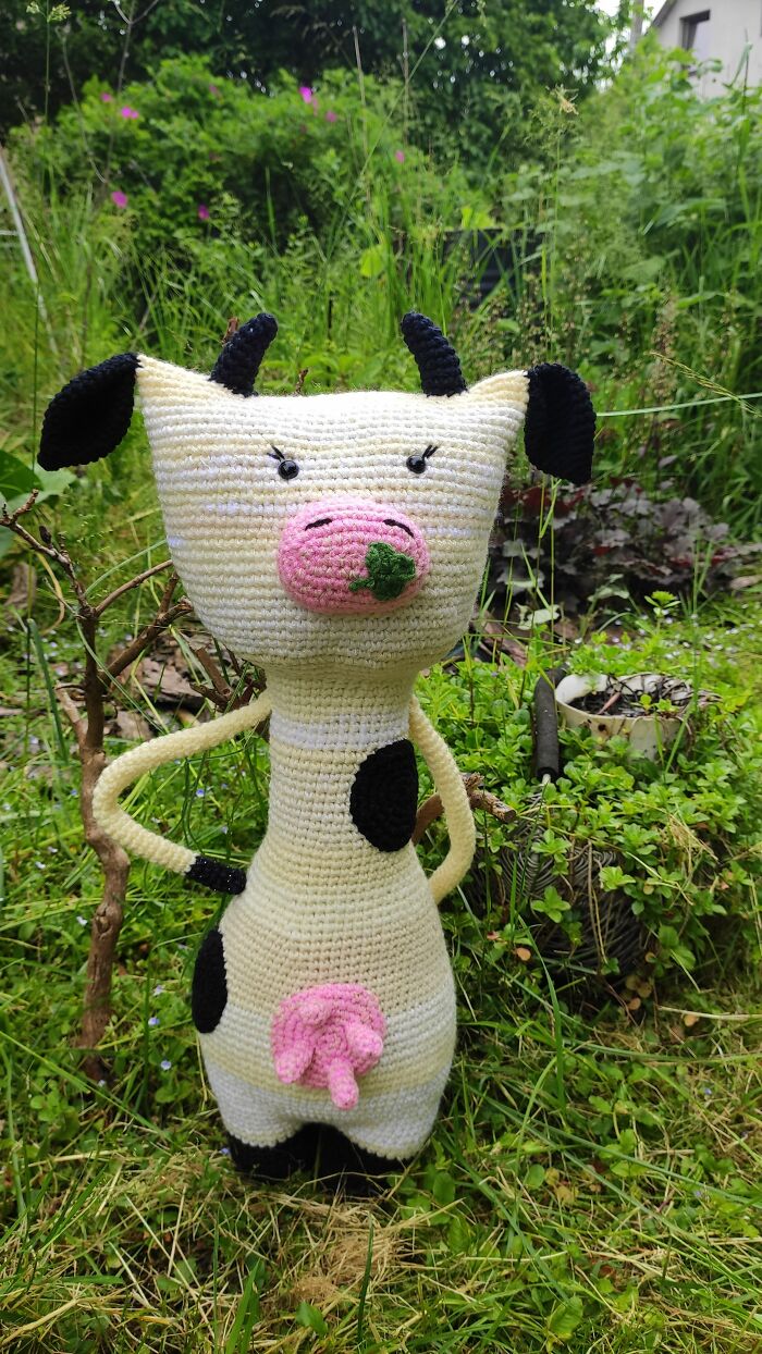 A white and black spotted crochet cow with a pink nose stands in the green grass.