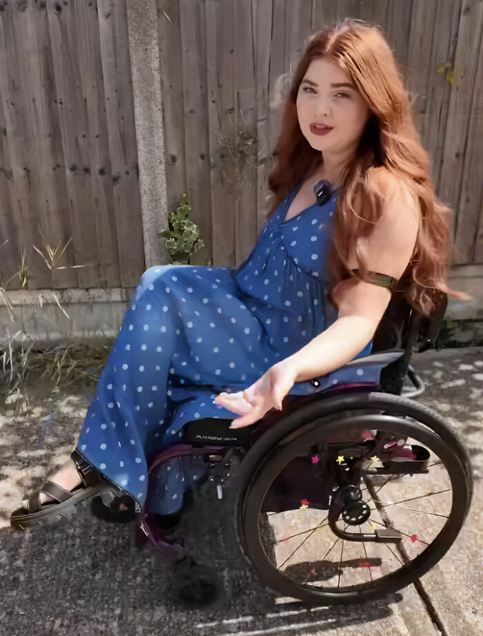 A young woman with long red hair wearing a blue dress while seated in a wheelchair mobility aid.