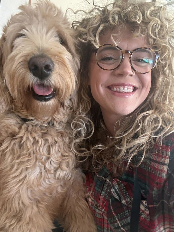 A smiling woman with curly hair and glasses next to a fluffy dog that looks like its owner.