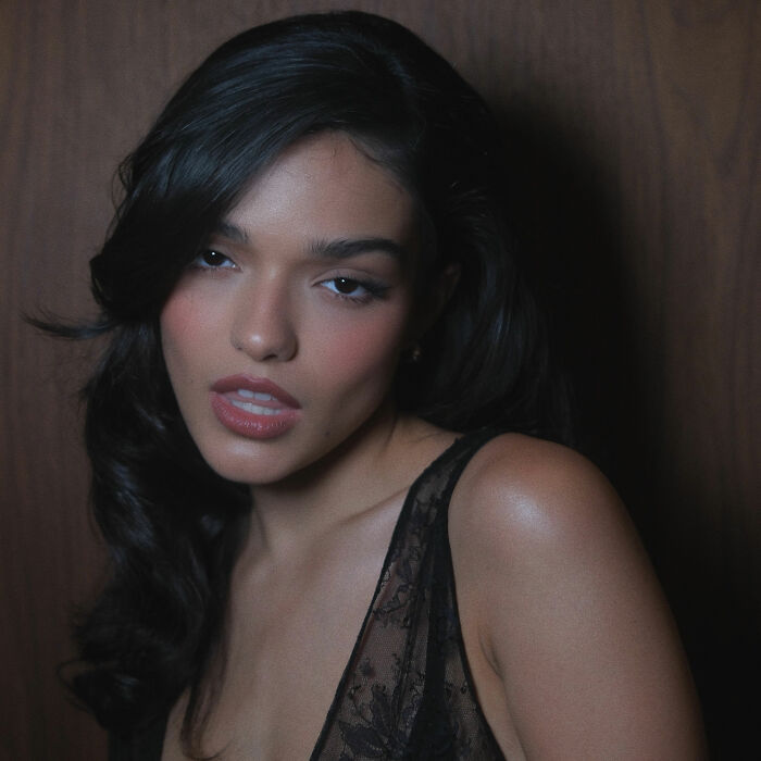 Rachel Zegler posing with slightly parted lips against a dark wooden wall.