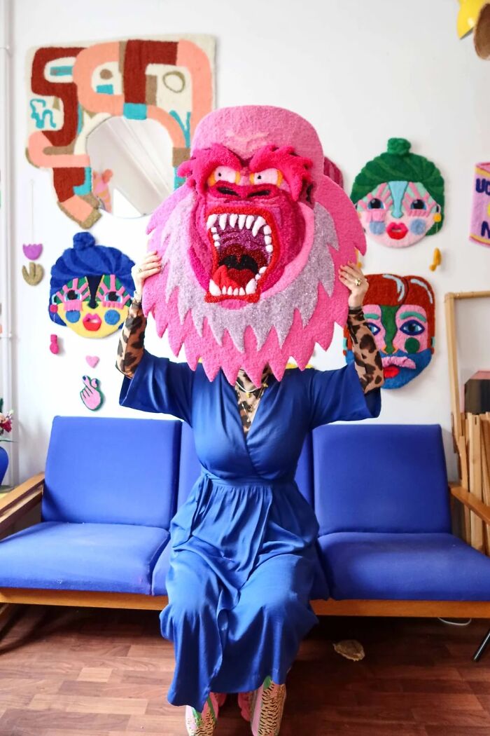 Artist Ina Dyreborg holding a pink roaring gorilla rug mask while seated on a blue sofa.