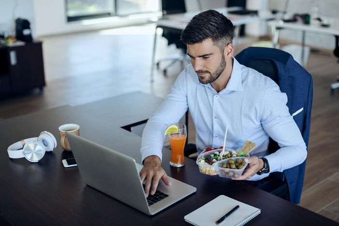 Man working at laptop holding container with chicken and sides for lunch Man working at laptop holding container with chicken and sides for lunch