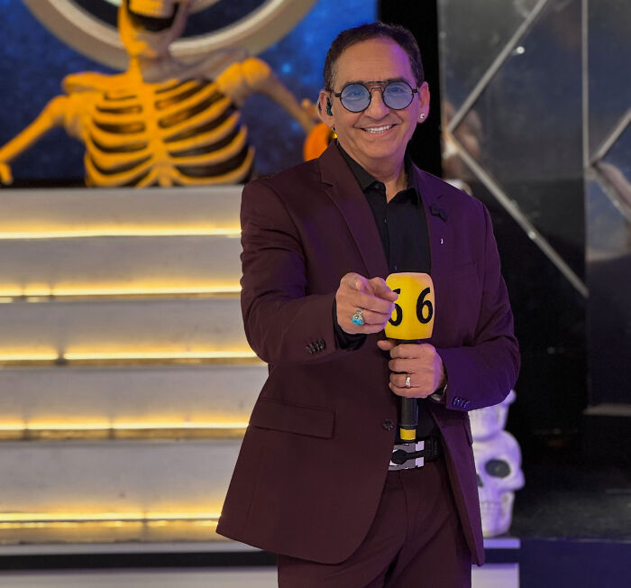 TV show host in a suit pointing forward while holding a microphone on stage.