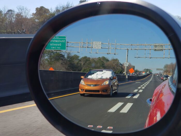 Absurd moment viewed in a side mirror showing a car driving with snow covering its windshield