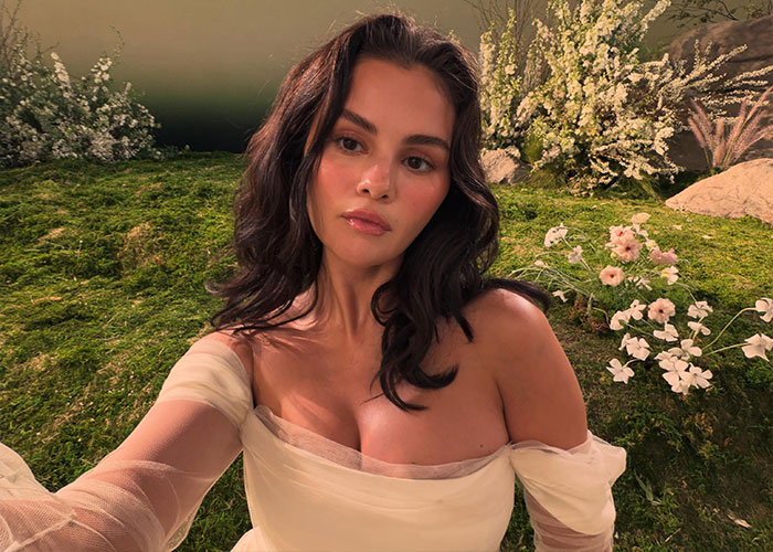 Selena Gomez taking a selfie in an off-the-shoulder white dress surrounded by flowers.