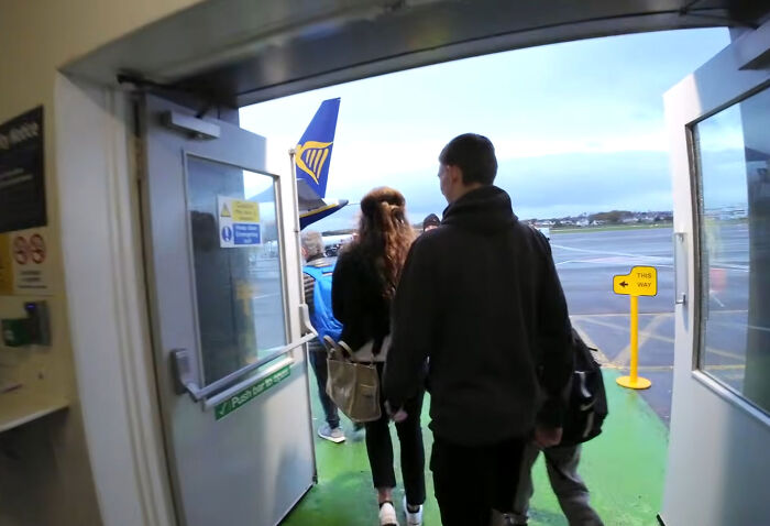 Passengers exiting an airport gate door onto the tarmac toward a waiting airplane.