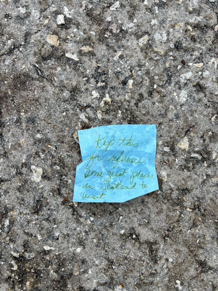A blue sticky note with handwritten text lying on the pavement, serving as a found paper note