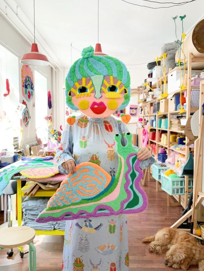 Artist Ina Dyreborg posing with her colorful handmade snail rug and character mask in her studio.