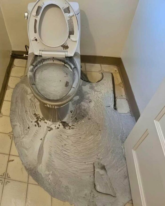 A disastrous DIY project where grey self leveling concrete has flooded a bathroom floor and splattered the toilet