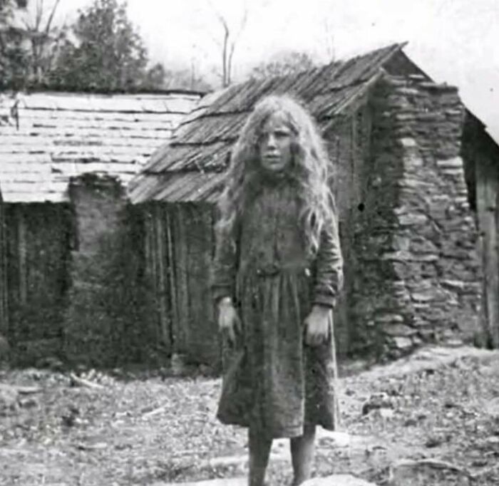 Black and white photo of a girl with long hair standing in front of rustic wooden and stone buildings