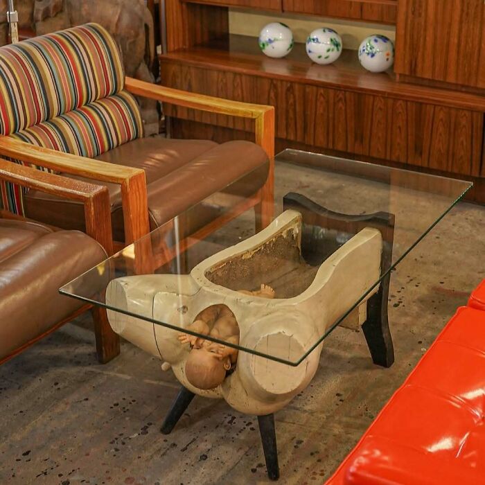 A weird glass coffee table supported by a creepy base shaped like a torso with a doll head protruding.