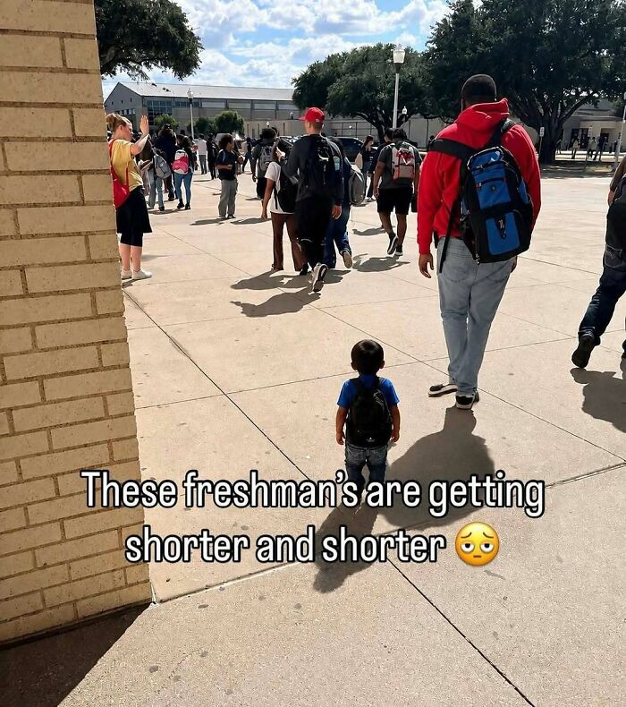 A toddler wearing a backpack walks among older students on a school walkway in hilarious pics.