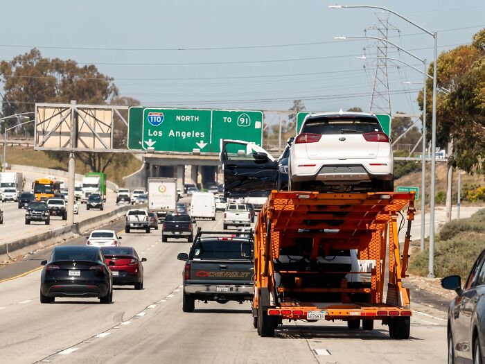 An orange car carrier truck driving on a highway, depicting towing gone wrong with a detached car door.